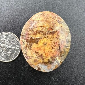 May include: A close-up of a polished, oval-shaped agate stone with a mix of brown, orange, and white colors. The stone has a unique, swirling pattern and a smooth, polished surface.