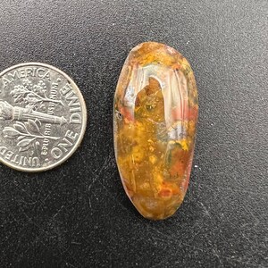 May include: A polished, oval-shaped gemstone with a mix of colors including yellow, orange, red, and clear. The gemstone is next to a U.S. dime coin for size comparison. The stone has a glossy, reflective surface.