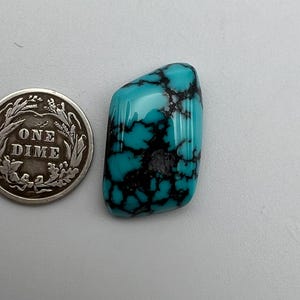 May include: A polished, rectangular turquoise gemstone with a vibrant cyan blue color and prominent black matrix veining, positioned next to a silver-colored United States one dime coin for scale, showing the text ONE DIME on its face.