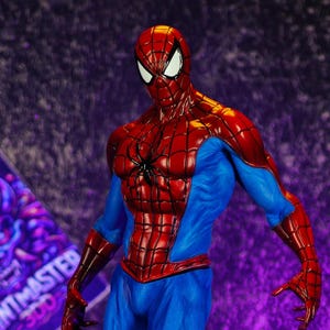 Puede incluir: Figura detallada de Spider-Man en una pose din&aacute;mica, luciendo su ic&oacute;nico traje rojo y azul. La figura se encuentra sobre una base texturizada, con un fondo p&uacute;rpura. Un letrero con el texto "PRINTMASTER 300" es visible.