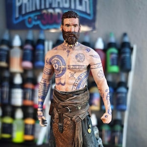 Puede incluir: Figura detallada de un hombre barbudo con tatuajes azules en el pecho y los brazos. Viste ropa marrón y negra con un delantal de piel. La figura está sobre una base rocosa, con el texto "PRINTMASTER 3D" visible en el fondo.