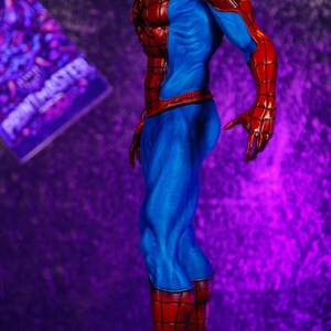 Puede incluir: Una figura detallada de Spider-Man en una pose din&aacute;mica. La figura es predominantemente roja y azul, con un patr&oacute;n de telara&ntilde;a texturizado en las secciones rojas. La base es una superficie rocosa y texturizada. El fondo es un degradado de p&uacute;rpura.