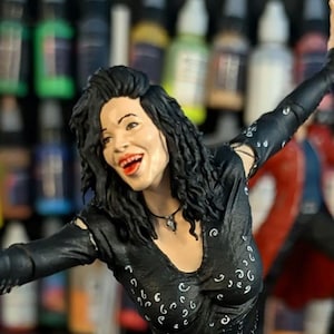 May include: A figurine of a woman with black hair and red lipstick, wearing a black dress with white detailing. The figurine is smiling with arms outstretched. The background includes paint bottles and the text "PRINTMASTER 300".