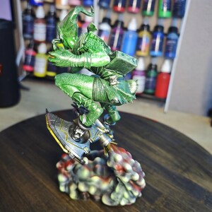 Puede incluir: Figura en miniatura detallada y pintada a mano de un duende verde montado en un dispositivo a reacci&oacute;n. La figura est&aacute; sobre una superficie de madera, con un estante de botes de pintura en el fondo. La figura es muy detallada.