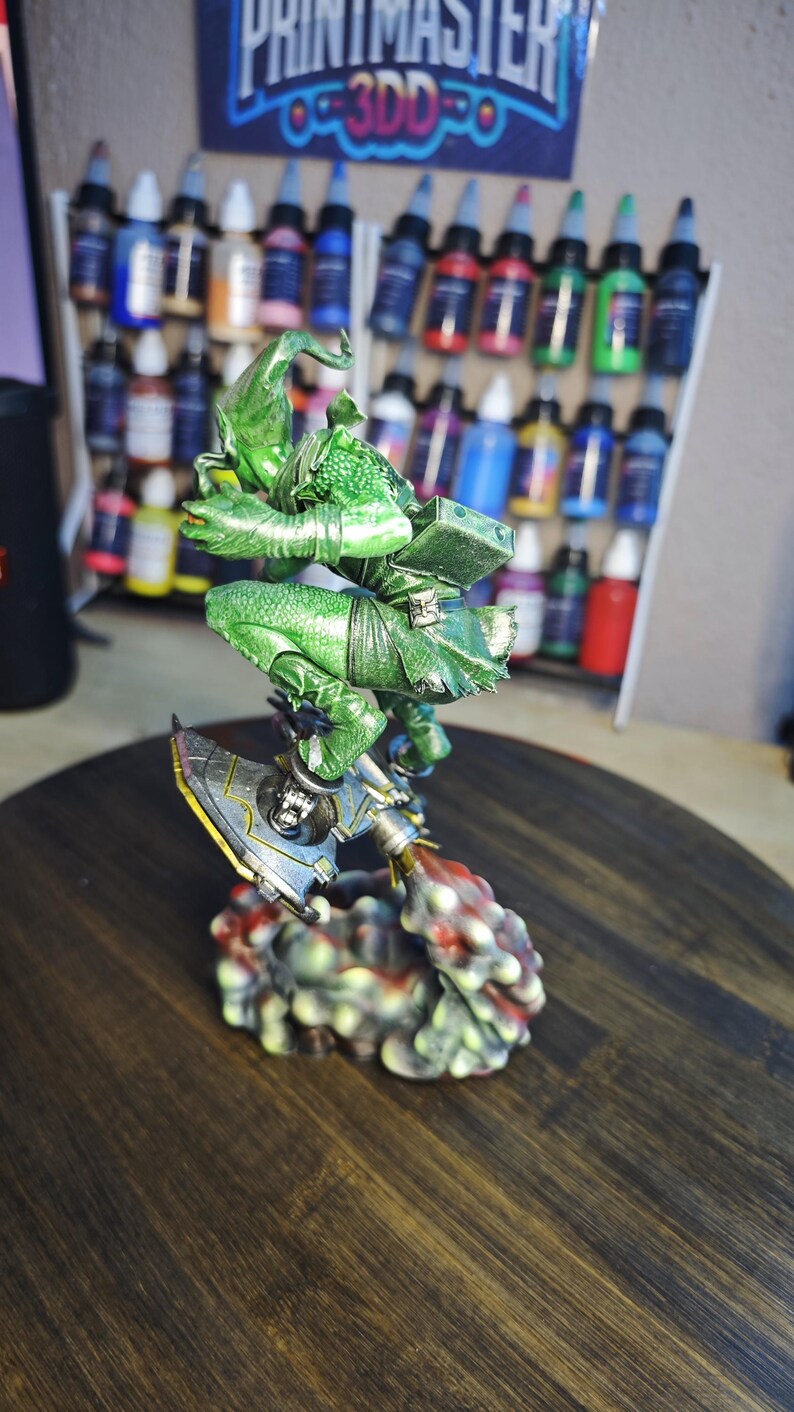 Puede incluir: Figura en miniatura detallada y pintada a mano de un duende verde montado en un dispositivo a reacci&oacute;n. La figura est&aacute; sobre una superficie de madera, con un estante de botes de pintura en el fondo. La figura es muy detallada.