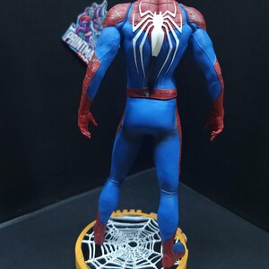 May include: A red and blue action figure of Spider-Man standing on a web-shaped base. The figure is posed with its back to the camera, showing the white spider symbol on its back.