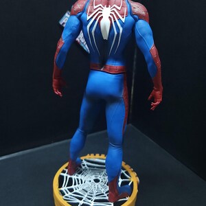 May include: A red and blue Spider-Man action figure with a white spider symbol on the back. The figure is standing on a round base with a white spider web design.