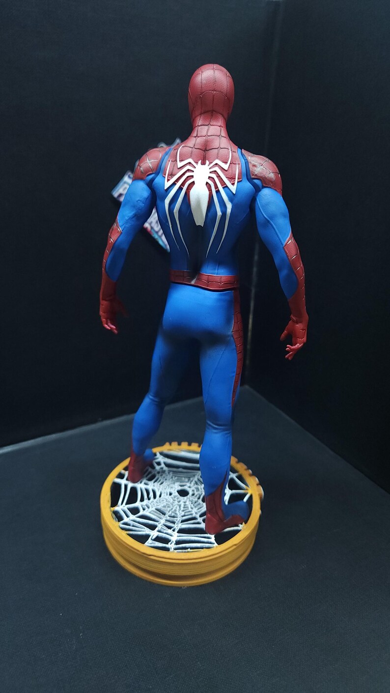 May include: A red and blue Spider-Man action figure with a white spider symbol on the back. The figure is standing on a round base with a white spider web design.