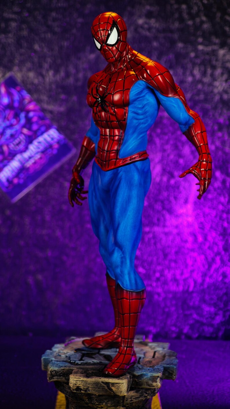 Puede incluir: Figura detallada de Spider-Man en una pose din&aacute;mica. La figura es predominantemente roja y azul, con un patr&oacute;n de telara&ntilde;a en las secciones rojas. La figura se alza sobre una base texturizada detallada, sobre un fondo p&uacute;rpura.