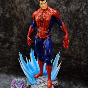 Op de afbeelding: Een gedetailleerd beeldje van Spider-Man in een dynamische pose, met zijn iconische rood-blauwe pak. Het beeldje staat op een rotsachtige basis met blauwe energie-effecten en heeft een zwart spinnenembleem op de borst. De basis bevat het "PRINTMASTER" logo.