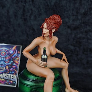 Può includere: Una statuina nuda di una donna con i capelli rossi, che tiene in mano una bottiglia scura con un tappo verde. È seduta su una base cilindrica verde. Sullo sfondo è visibile una scatola colorata con la scritta "MASTER GOD".
