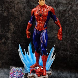 Op de afbeelding: Een gedetailleerd beeldje van Spider-Man in een dynamische pose, met een rood en blauw kostuum en een zwart spinnenembleem. De figuur staat op een rotsachtige basis met blauwe energie-effecten. Een losgemaakt masker en hand zijn ook aanwezig. De basis bevat een "PRINTMASTER" sticker.