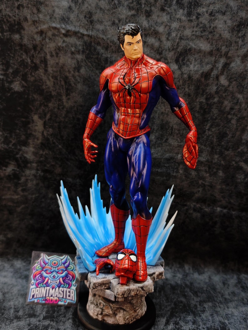 Op de afbeelding: Een gedetailleerd beeldje van Spider-Man in een dynamische pose, met een rood en blauw kostuum en een zwart spinnenembleem. De figuur staat op een rotsachtige basis met blauwe energie-effecten. Een losgemaakt masker en hand zijn ook aanwezig. De basis bevat een "PRINTMASTER" sticker.