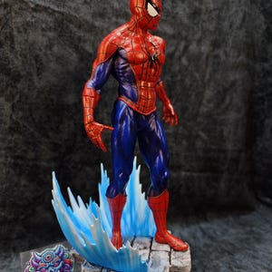 Op de afbeelding: Gedetailleerd beeldje van Spider-Man in een dynamische pose, met zijn iconische rood-blauwe pak. Het beeldje staat op een rotsachtige basis met een gestileerd watereffect. Het Printmaster-logo is zichtbaar.