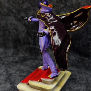 Pu&ograve; includere: Statuetta di un personaggio in un completo viola e nero con dettagli dorati, un mantello e una maschera. La figura &egrave; su una base a pi&ugrave; livelli con un tappeto rosso. Lo sfondo &egrave; grigio scuro.