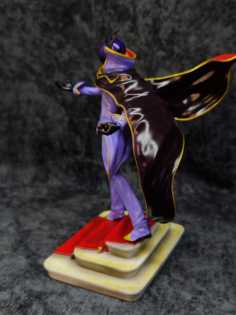 Pu&ograve; includere: Statuetta di un personaggio in un completo viola e nero con dettagli dorati, un mantello e una maschera. La figura &egrave; su una base a pi&ugrave; livelli con un tappeto rosso. Lo sfondo &egrave; grigio scuro.