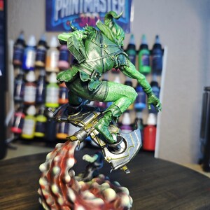Puede incluir: Una figura verde de un personaje de fantas&iacute;a en una pose din&aacute;mica, con un arma y una base en forma de llama. La figura est&aacute; detallada con armadura y se encuentra frente a un fondo de botes de pintura y un p&oacute;ster que dice "PRINTMASTER".