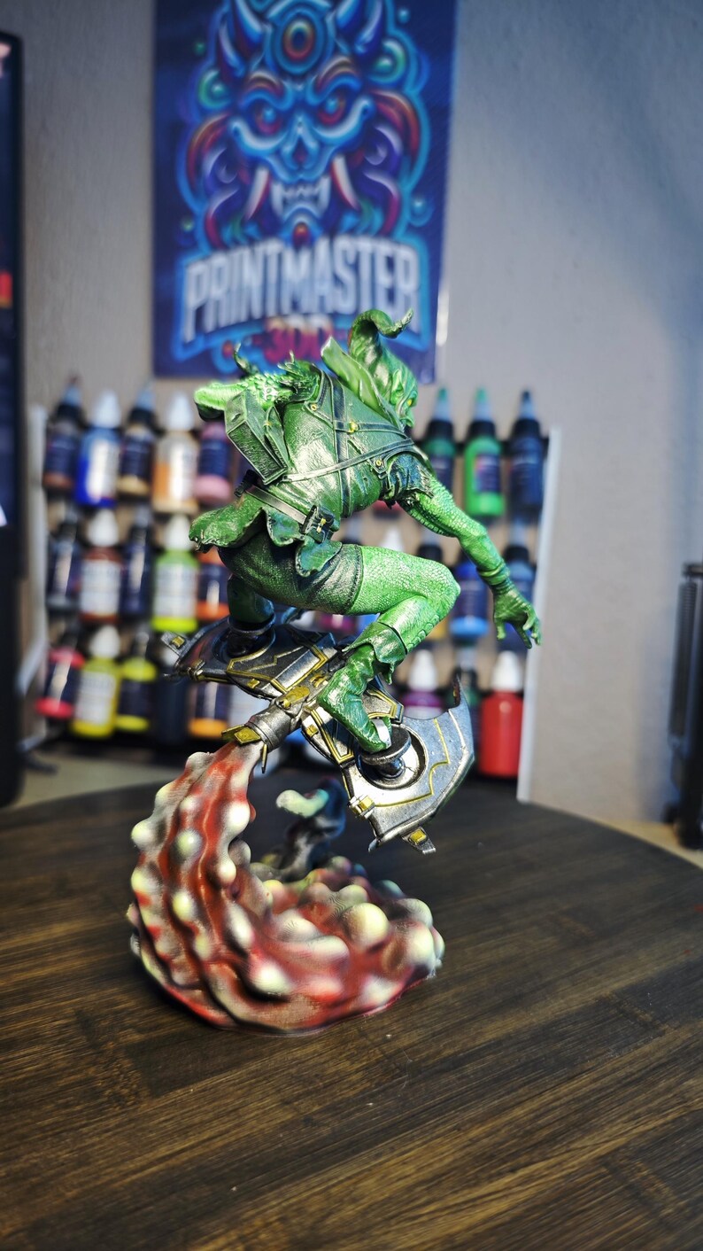 Puede incluir: Una figura verde de un personaje de fantas&iacute;a en una pose din&aacute;mica, con un arma y una base en forma de llama. La figura est&aacute; detallada con armadura y se encuentra frente a un fondo de botes de pintura y un p&oacute;ster que dice "PRINTMASTER".