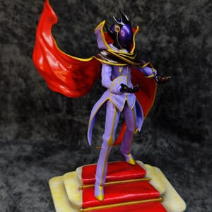 Pu&ograve; includere: Statuetta di un personaggio in un completo viola e oro con un mantello rosso. La figura &egrave; in piedi su una piattaforma con moquette rossa con finiture dorate. Il personaggio ha un elmo con una visiera viola.