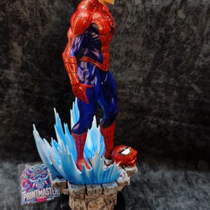 Op de afbeelding: Een gedetailleerd beeldje van Spider-Man in een dynamische pose, gekleed in zijn iconische rood-blauwe pak. Het beeld staat op een voetstuk met waterachtige blauwe accenten en een los masker. Het beeld is ongeveer 30 cm hoog.