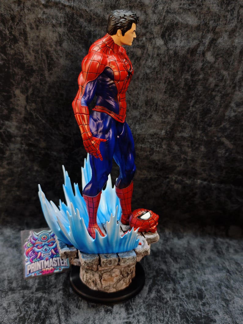 Op de afbeelding: Een gedetailleerd beeldje van Spider-Man in een dynamische pose, gekleed in zijn iconische rood-blauwe pak. Het beeld staat op een voetstuk met waterachtige blauwe accenten en een los masker. Het beeld is ongeveer 30 cm hoog.