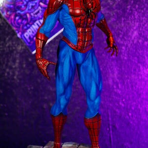 Puede incluir: Figura detallada de Spider-Man con su cl&aacute;sico traje rojo y azul. La figura est&aacute; posada din&aacute;micamente sobre una base texturizada, con un fondo p&uacute;rpura y azul. La figura est&aacute; en una pose din&aacute;mica, con los brazos extendidos.