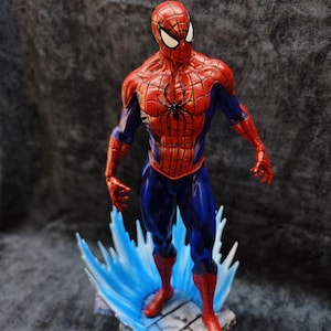 Op de afbeelding: Een gedetailleerd beeldje van Spider-Man in een dynamische pose. De figuur is overwegend rood en blauw, met een zwart spinnenembleem op de borst. Het staat op een voetstuk met blauwe watereffecten, wat het iconische kostuum van de superheld laat zien.