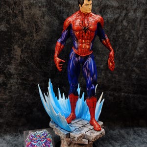 Op de afbeelding: Gedetailleerd beeldje van Spider-Man in een dynamische pose, gekleed in zijn iconische rood-blauwe pak. Het beeld staat op een stenen voet met blauwe watereffecten. De basis heeft een zwarte cirkelvormige standaard. Het beeld heeft een zwart spinnenembleem op de borst.