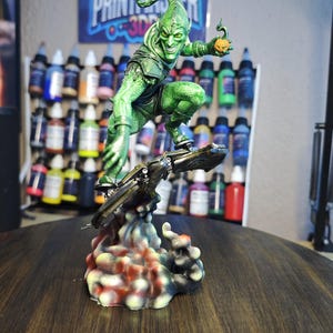 Puede incluir: Una detallada figura del Duende Verde, un villano de c&oacute;mic, montado en un planeador. La figura es predominantemente verde con una bomba de calabaza en la mano. La base es multicolor, y el fondo presenta botes de pintura y un p&oacute;ster con el texto "PRINTMASTER".