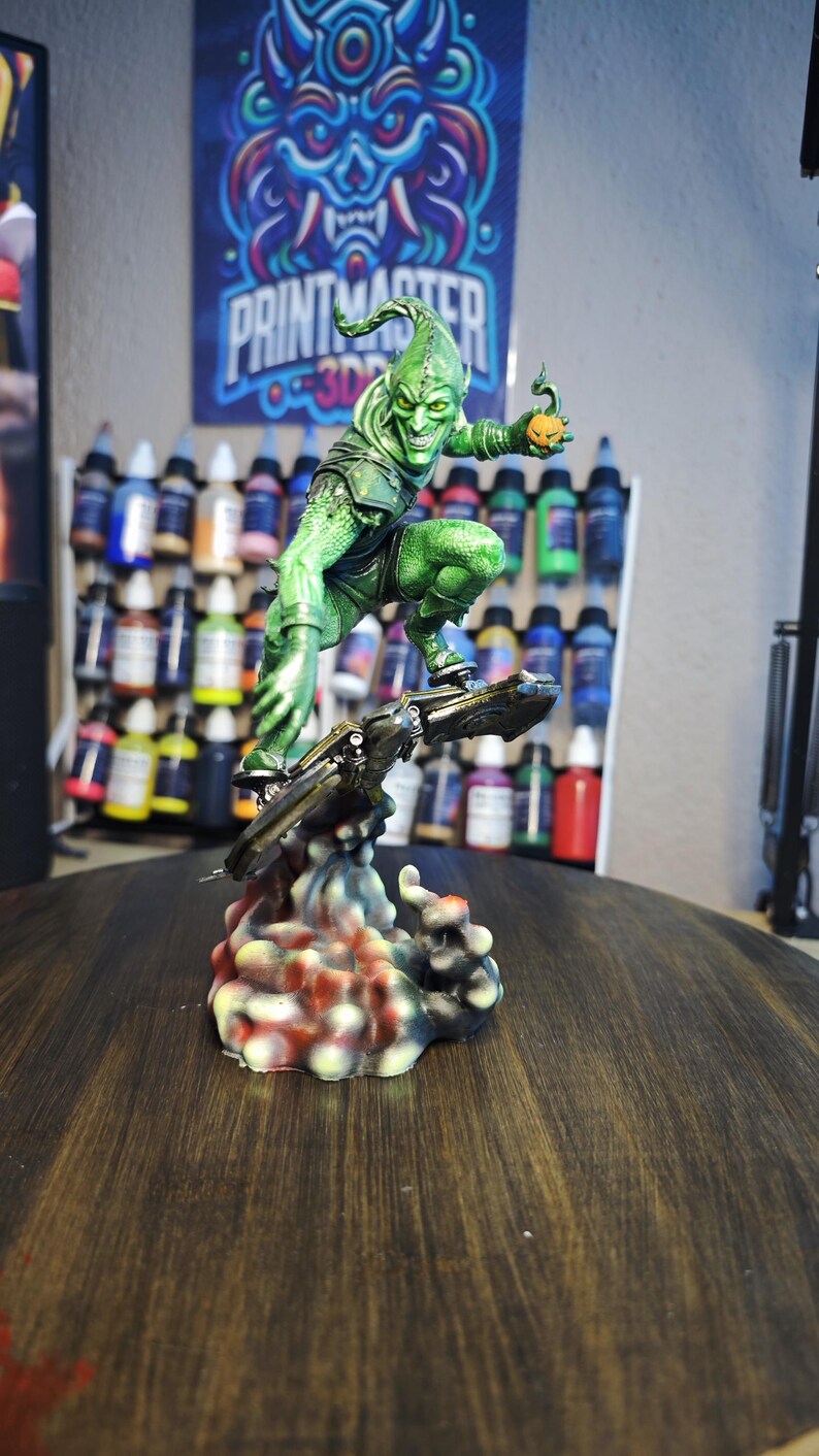 Puede incluir: Una detallada figura del Duende Verde, un villano de c&oacute;mic, montado en un planeador. La figura es predominantemente verde con una bomba de calabaza en la mano. La base es multicolor, y el fondo presenta botes de pintura y un p&oacute;ster con el texto "PRINTMASTER".