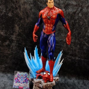 Op de afbeelding: Gedetailleerd beeldje van Spider-Man in een dynamische pose, gekleed in zijn kenmerkende rood-blauwe pak. Het beeldje staat op een rotsachtige basis met blauwe energie-effecten. Een losgemaakt masker ligt in de buurt. Het Printmaster-logo is zichtbaar.
