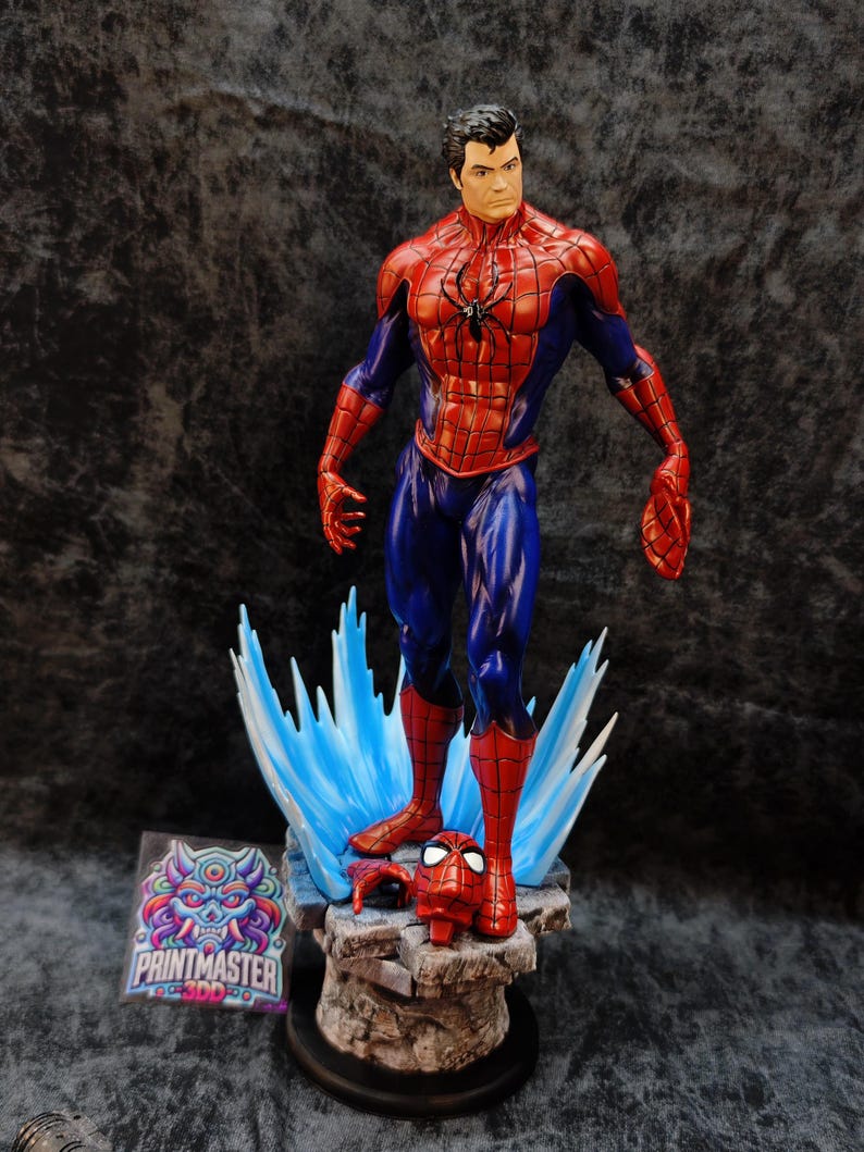 Op de afbeelding: Gedetailleerd beeldje van Spider-Man in een dynamische pose, gekleed in zijn kenmerkende rood-blauwe pak. Het beeldje staat op een rotsachtige basis met blauwe energie-effecten. Een losgemaakt masker ligt in de buurt. Het Printmaster-logo is zichtbaar.