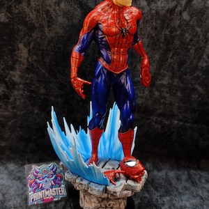 Op de afbeelding: Gedetailleerd beeldje van Spider-Man in een dynamische pose, gekleed in een rood en blauw pak. Het beeld staat op een rotsachtige basis met blauwe watereffecten, en een losgemaakt masker is in de buurt. Zwart spinnenembleem op de borst.