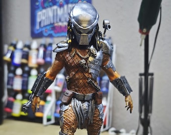 May include: A detailed Predator action figure, with a silver helmet, intricate armour, and a brown and orange body. The figure has a weapon on its shoulder and is posed in a dynamic stance. The background includes paint bottles and a sign that says PRINTMAKER.