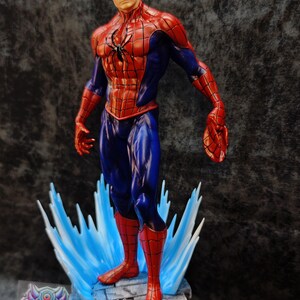 Op de afbeelding: Een gedetailleerd beeldje van Spider-Man in een dynamische pose, met zijn iconische rood-blauwe pak. Het beeld staat op een voetstuk met blauwe waterachtige accenten. Het beeld heeft een zwart spinnenembleem op de borst.