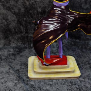 Pu&ograve; includere: Statuetta di un personaggio con un mantello viola e nero con finiture dorate. La figura &egrave; su una base a pi&ugrave; livelli color crema con un tappeto rosso. Il personaggio ha un elmo viola e tiene in mano un piccolo oggetto.