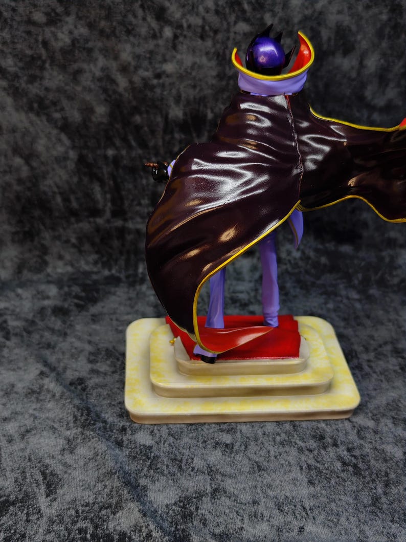 Pu&ograve; includere: Statuetta di un personaggio con un mantello viola e nero con finiture dorate. La figura &egrave; su una base a pi&ugrave; livelli color crema con un tappeto rosso. Il personaggio ha un elmo viola e tiene in mano un piccolo oggetto.