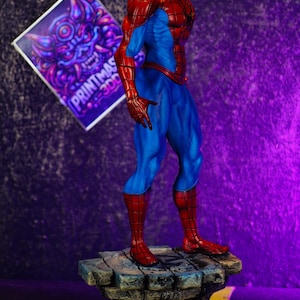Puede incluir: Una figura detallada de Spider-Man en una pose cl&aacute;sica, con un traje rojo y azul. La figura est&aacute; sobre una base de piedra. El fondo es un degradado de p&uacute;rpura y negro. Un letrero con el texto "PRINTMAS 300" est&aacute; en el fondo.