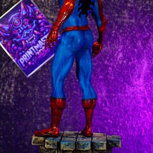 Puede incluir: Figura de Spider-Man impresa en 3D, vista desde atr&aacute;s, sobre una base de piedra. La figura lleva un traje rojo y azul con un emblema de ara&ntilde;a negro. Un gr&aacute;fico morado y blanco est&aacute; en el fondo.