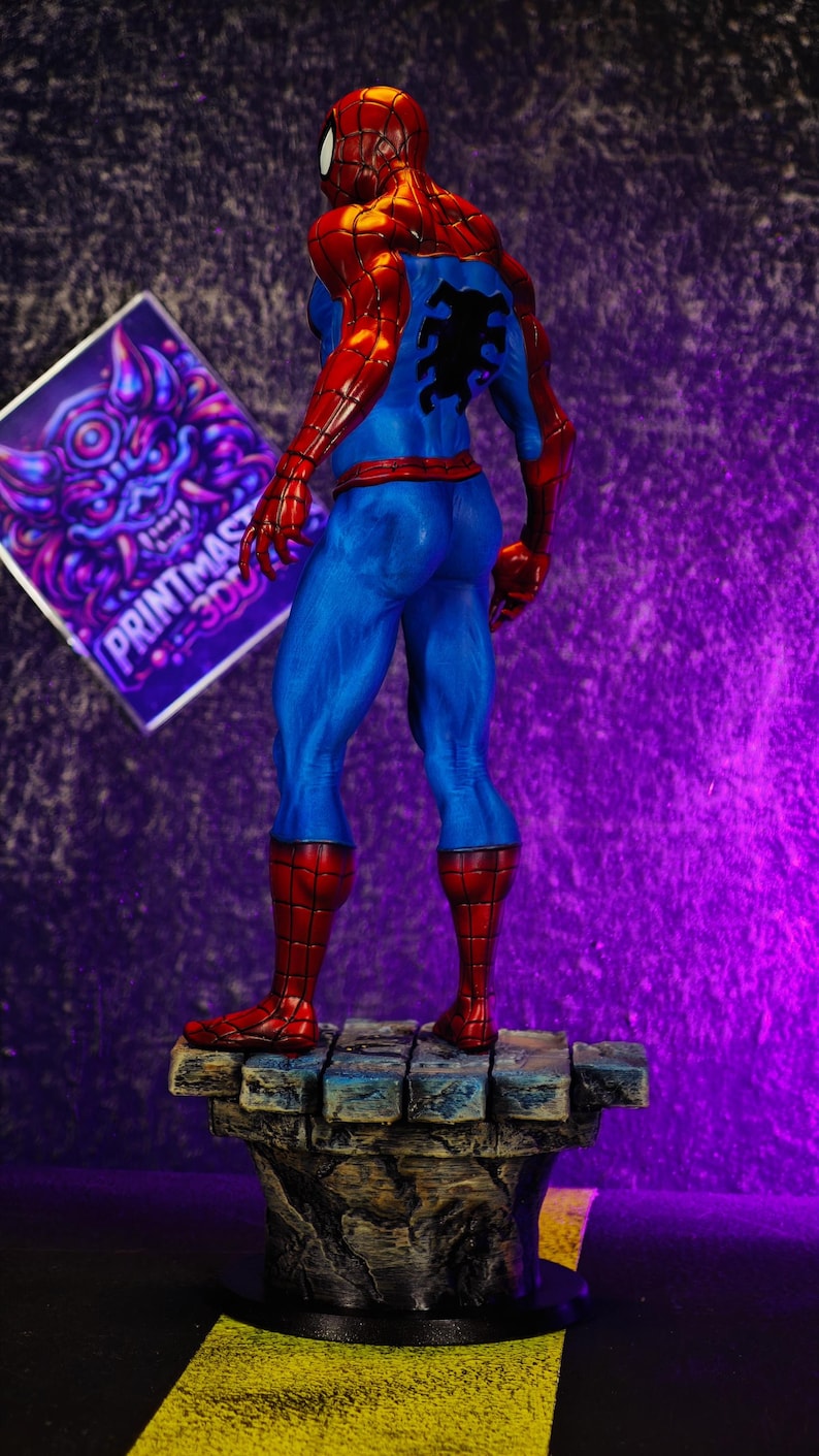 Puede incluir: Figura de Spider-Man impresa en 3D, vista desde atr&aacute;s, sobre una base de piedra. La figura lleva un traje rojo y azul con un emblema de ara&ntilde;a negro. Un gr&aacute;fico morado y blanco est&aacute; en el fondo.
