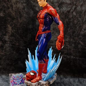 Op de afbeelding: Gedetailleerd beeldje van Spider-Man in zijn iconische rood-blauwe pak, in een dynamische pose. Het beeld staat op een rotsachtige basis met blauwe watereffecten en bevat een los masker. Het beeldje heeft een gedetailleerd gezicht.