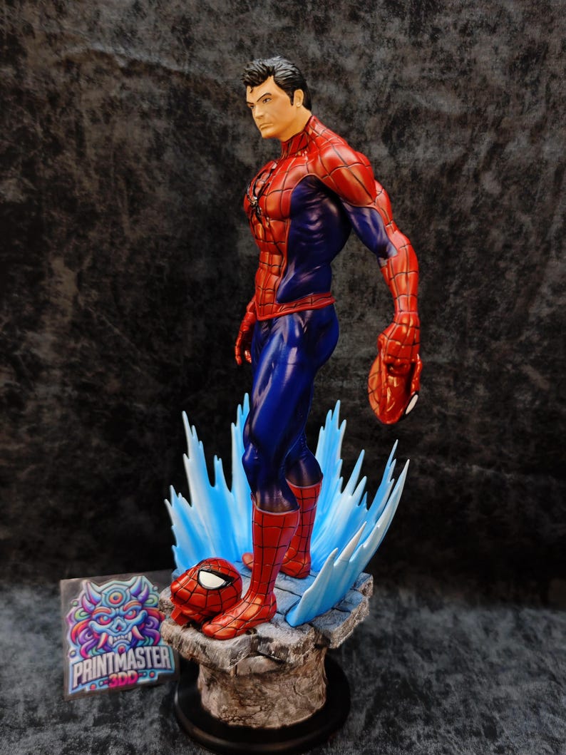 Op de afbeelding: Gedetailleerd beeldje van Spider-Man in zijn iconische rood-blauwe pak, in een dynamische pose. Het beeld staat op een rotsachtige basis met blauwe watereffecten en bevat een los masker. Het beeldje heeft een gedetailleerd gezicht.