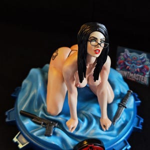 May include: A figurine of a woman kneeling on a blue surface, wearing glasses and black hair. A handgun and a knife are placed on the surface. The figurine is on a blue and black base with a red emblem.