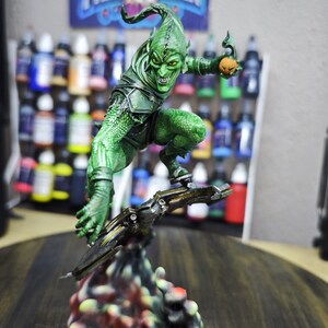 Puede incluir: Una detallada figura del Duende Verde, un villano de c&oacute;mic, en una pose din&aacute;mica. La figura es predominantemente verde con una expresi&oacute;n amenazante, sosteniendo una bomba de calabaza. La base es un dise&ntilde;o colorido y texturizado. Un p&oacute;ster con el texto "PRINTMASTER" est&aacute; en el fondo.