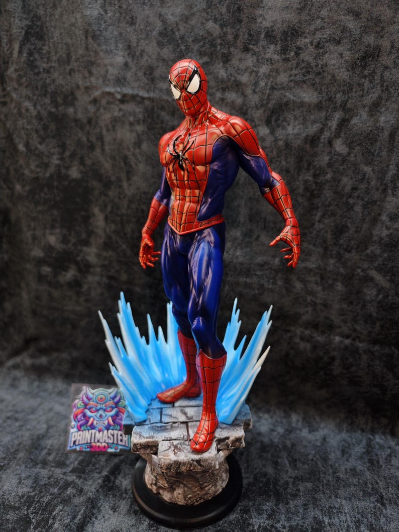 Op de afbeelding: Een gedetailleerd beeldje van Spider-Man in een dynamische pose, gekleed in zijn kenmerkende rood-blauwe pak. Het beeldje staat op een stenen voet met blauwe energie-effecten, met ingewikkelde details en levendige kleuren. De voet heeft de tekst "PRINTMASTER".