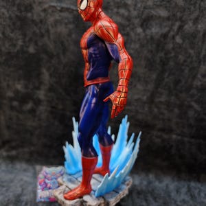 Op de afbeelding: Een gedetailleerd beeldje van Spider-Man in een dynamische pose, met zijn iconische rood-blauwe pak. Het beeldje staat op een rotsachtige basis met blauwe watereffecten. De figuur is ongeveer 20 cm hoog.