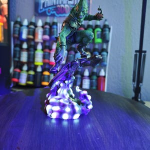Puede incluir: Una detallada figura del Duende Verde, un personaje de c&oacute;mic, en una pose din&aacute;mica sobre una base p&uacute;rpura brillante. La figura es verde con una expresi&oacute;n amenazante. El fondo incluye botes de pintura y un p&oacute;ster con el texto "PRINTMASTERS".