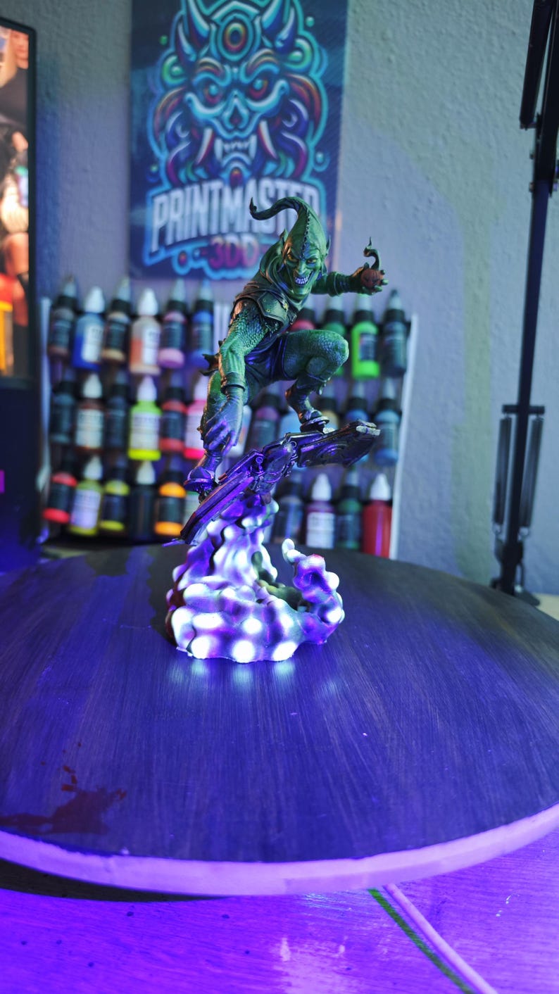 Puede incluir: Una detallada figura del Duende Verde, un personaje de c&oacute;mic, en una pose din&aacute;mica sobre una base p&uacute;rpura brillante. La figura es verde con una expresi&oacute;n amenazante. El fondo incluye botes de pintura y un p&oacute;ster con el texto "PRINTMASTERS".