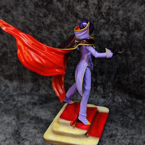 Pu&ograve; includere: Una statuetta di un personaggio in un completo viola e giallo con un mantello rosso svolazzante. La figura &egrave; in piedi su una base a pi&ugrave; livelli con un design a tappeto rosso. Il personaggio ha una maschera e un cappuccio.