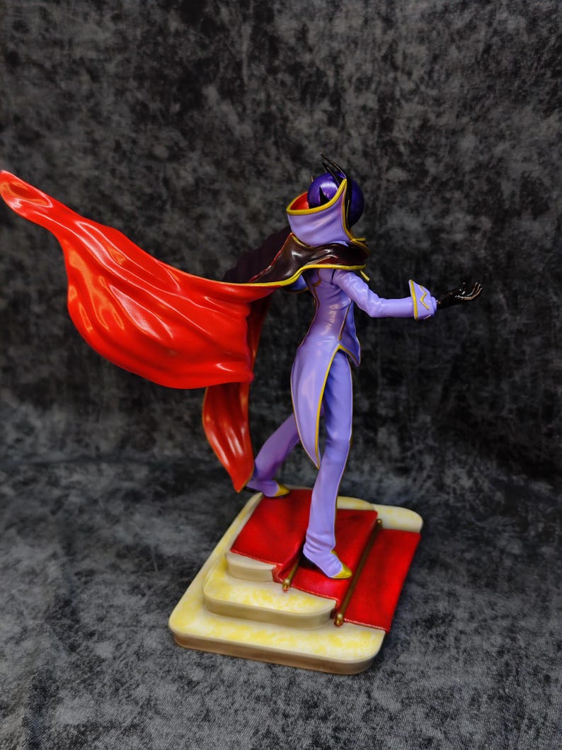 Pu&ograve; includere: Una statuetta di un personaggio in un completo viola e giallo con un mantello rosso svolazzante. La figura &egrave; in piedi su una base a pi&ugrave; livelli con un design a tappeto rosso. Il personaggio ha una maschera e un cappuccio.